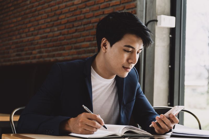 young asian man taking notes and looking at his phone